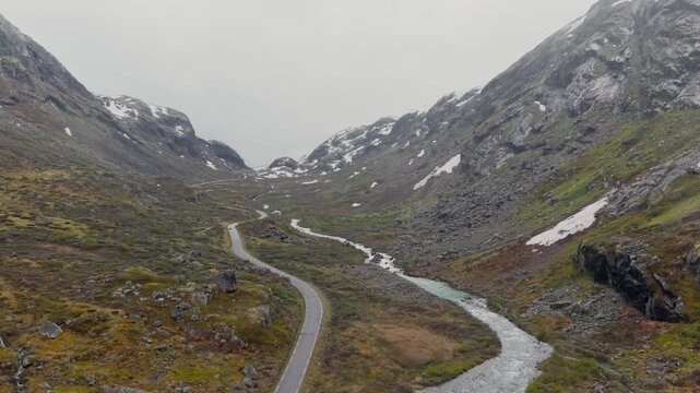 Drohnenflug &uuml;ber Stra&szlig;e neben Fluss in Skandinavien, umgeben von verschneiten Berggipfeln, mosige Landschaft bei nebligem Wetter. RAW unbearbeitet