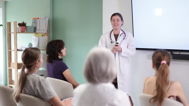 Positive young girl medical doctor dressed in white medical coat with stethoscope standing and talking in auditorium where sitting medical student different ages