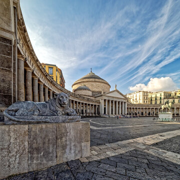 A majestic marble lion rests in Piazza del Plebiscito, Naples. Wide-angle view featuring the historic cobblestone plaza, neoclassical colonnade, and the Basilica dome under a dramatic sky.