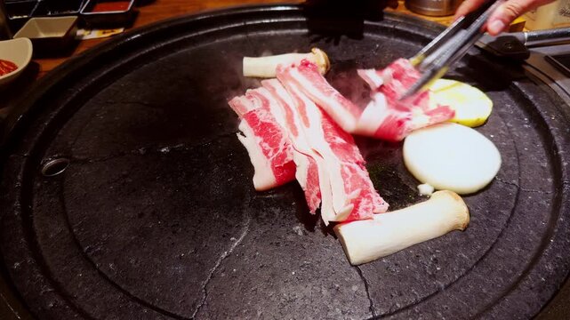Thin Slices of Raw Beef Laid on Hot Grill Beside Mushrooms and Onion While Tongs Arrange Meat Under Bright Warm Light