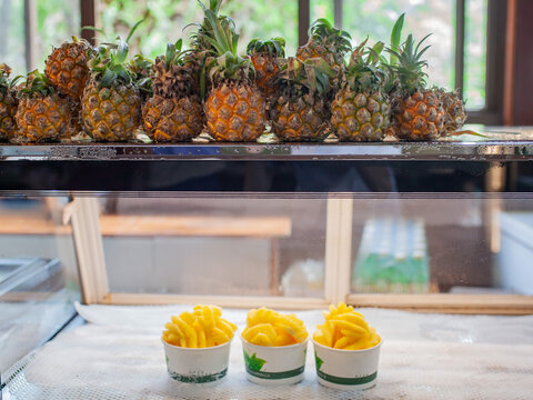 Fresh whole pineapples on top shelf and three paper cups with sliced pineapple on market counter. Tropical street-food snack concept. Freshness, healthy snacking, summer refreshment and takeaway.