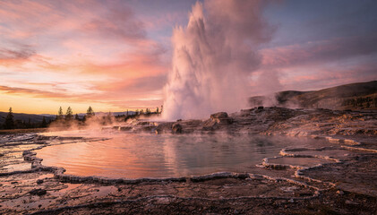 Fototapeta premium Dramatic Volcanic Geyser Erupts Against a Colorful Sunset Sky, with Steam Rising from Hot Springs and Rugged Geothermal Landscape Details