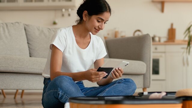 Smiling Hindu Indian woman at home preparing for trip vacation travel female Arabian girl browsing mobile phone ordering flight chatting booking hotel packing clothes suitcase tickets passport indoors