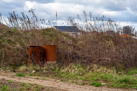 Two old rusty metal barrels abandoned on a wasteland near a construction site, environmental pollution in rural area, residential houses and wind turbines in the background under cloudy sky.