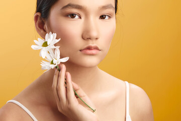 Fototapeta premium asian woman with delicate features holds white daisies near her face, posing against a warm backdrop, conveying natural beauty, calm confidence, and graceful simplicity.