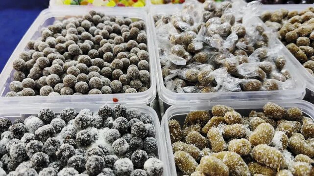 Tracking shot of multiple containers filled with sugar-coated tamarind candies and coated fruit treats, showcasing varied textures and colors in a vibrant Indian street market display!