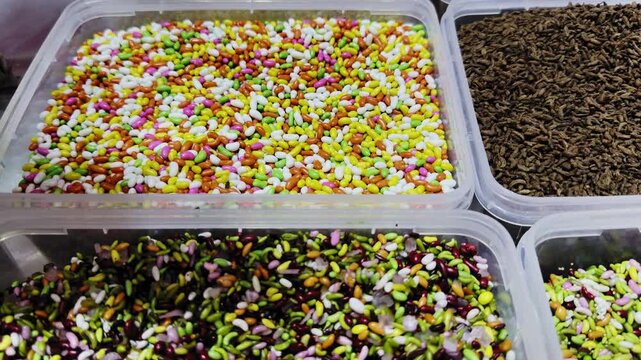 Tracking shot of multiple containers filled with colorful mouth freshener blends, featuring fennel seeds, sugar-coated saunf, and assorted mukhwas varieties in a vibrant market display!