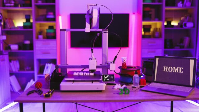 A 3D printer is actively creating the word HOME in white letters on its build plate, surrounded by filament spools and tools
