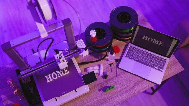 A 3D printer is actively creating the word HOME with white filament while a laptop displays the same word on its screen, surrounded by spools of filament and tools