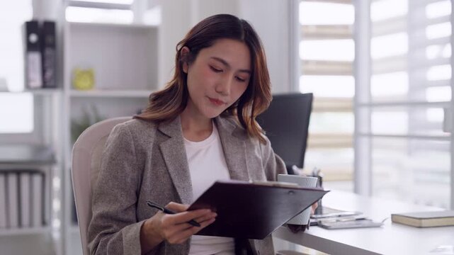 Asian adult business professional woman drinks coffee studies documents at desk modern office workspace planning strategy and reviewing reports