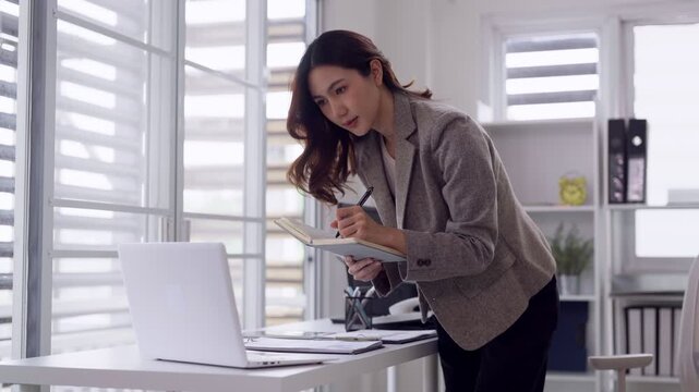 Asian adult business professional woman writing notes laptop computer modern office setting business strategy planning focused work environment
