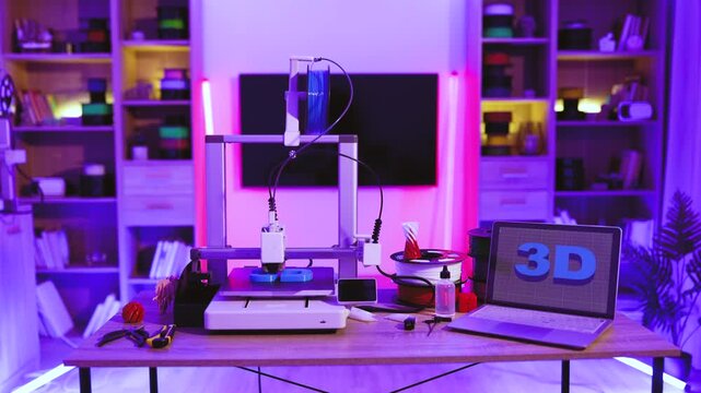 A 3D printer is actively creating an object on a desk, with a laptop displaying 3D graphics and various filament spools nearby