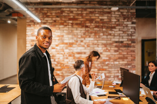 Man stands in office with papers while colleagues work at desks in a modern workplace environment