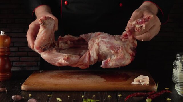 Chef in black uniform holds raw meat rabbit while preparing it on wooden cutting board with spices and kitchen tools visible in the background