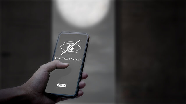 A person's hand holding a smartphone displaying a sensitive content warning message with a hidden eye icon, representing internet censorship and online safety