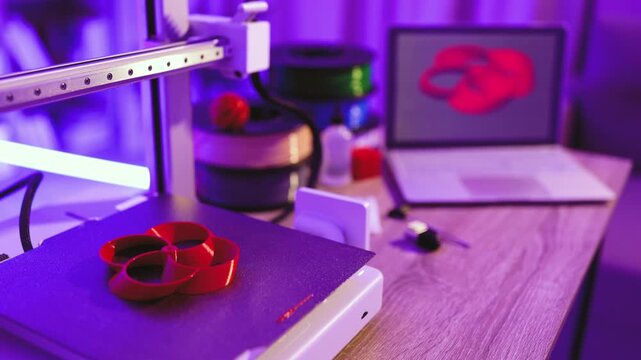 A 3D printer is actively creating a red object on its build plate, with filament spools and a laptop displaying the design in the background under purple lighting