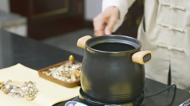 Herbalist apothecary medicine doctor hands adding herbal ingredients to an enamel steaming pot to electric heating decoct herbal medicine cooker kettle. Natural remedy alternative healthcare concept