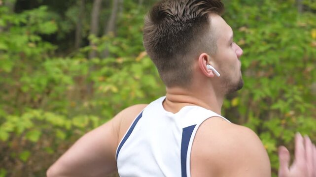 Athletic sporty guy with wireless headphones sprinting fast along trail near forest. Young strong sportsman jogging outdoor at early autumn. Handsome man running at nature. Healthy active lifestyle