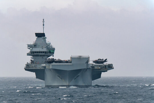 Royal Navy aircraft carrier HMS Prince of Wales sailing in Tokyo Bay, Japan, with F-35B fighter jets on deck, rough and cloudy weather.