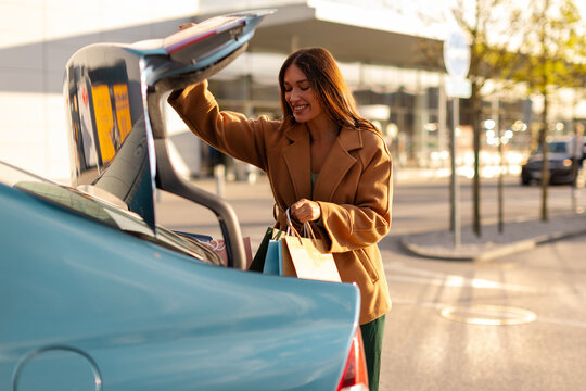 Fashionable lady standing on parking lot near car trunk and putting paper shopping bags into it, had fun on shopping spree at shopping center