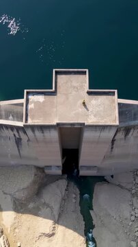 Vertical video: Hovering drone filming person in hi-vis jacket on dam checking outlet for low water