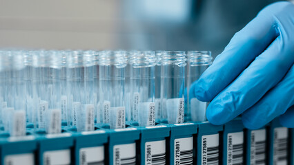 Laboratory technician wearing blue gloves handling test tubes in a laboratory setting, with organized racks and clear labels visible in the background © wedmoments.stock