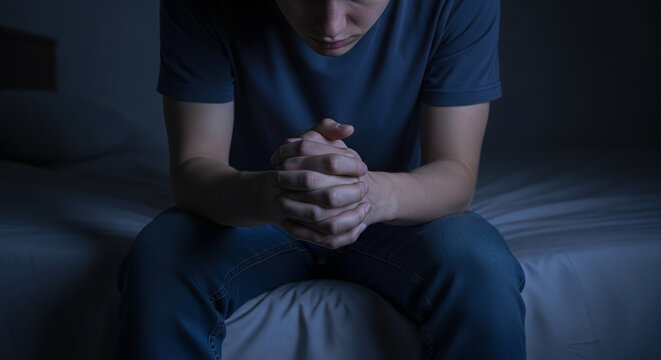A young man sits on the edge of his bed in a dimly lit room, his hands clasped together in thought or prayer.