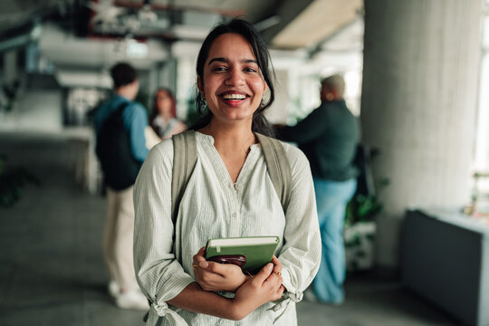 Happy indian female student smiling, embracing education and university life, holding book on college campus