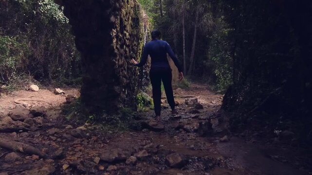 Mujer caminando con cuidado entre piedras y agua en un bosque frondoso con un acueducto