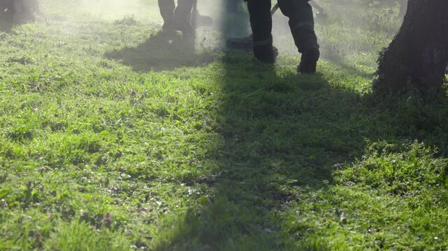 Lower body shot of workers walking and using brush cutters, moving from shadow into bright sunlight, clearing a visible path through the grass.