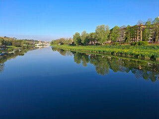 Obraz premium Beautiful morning view of a calm river reflecting the blue sky, green bank, and city architecture. Peaceful European town landscape during spring or summer.