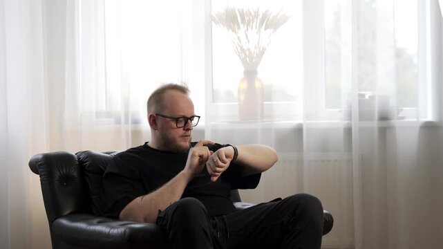 A man seated in a cozy room explores his smart watch, light streaming through sheer curtains. The setting is calm and modern, showcasing the intersection of technology and relaxation.