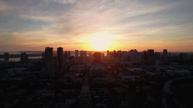 Drone flying forward toward the downtown San Diego skyline at sunset with glowing sunlight behind the buildings.