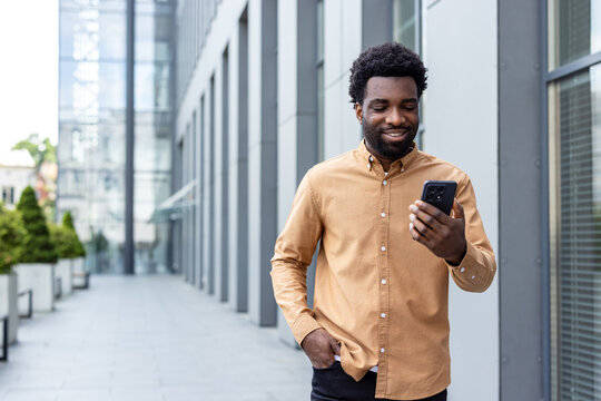 African person smiling while engaging with a mobile device outside a modern office building, representing communication, technology, and connection in a busy city setting