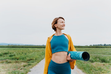 Obraz premium Beautiful fit woman holding yoga mat outdoors. Smiling female with exercise mat in green nature park