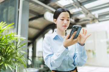 Fototapeta premium A young woman in a blue shirt uses her smartphone indoors, standing near green plants and modern architecture.