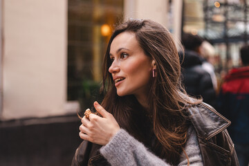 Side View Profile of a Beautiful Woman Looking Away with Curious and Engaged Expression. Wearing a Grey Mohair Sweater, Brown Leather Jacket, and Pink Earrings on a Blurred Urban Lifestyle Background © zvkate