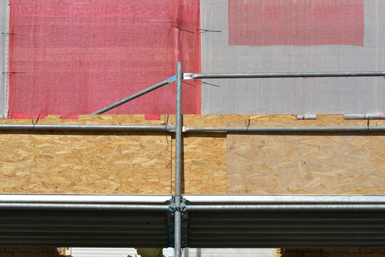 A detail view of metal scaffolding poles and connections securing oriented strand board panels and layered red and grey mesh on a building facade