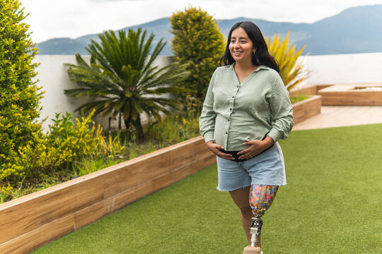 Mujer embarazada con pierna prot&eacute;sica abrazando la maternidad caminando por una terraza
