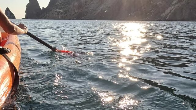 Kayak paddling ocean person rows orange boat on calm water near rocky cliffs at golden hour sunrise or sunset