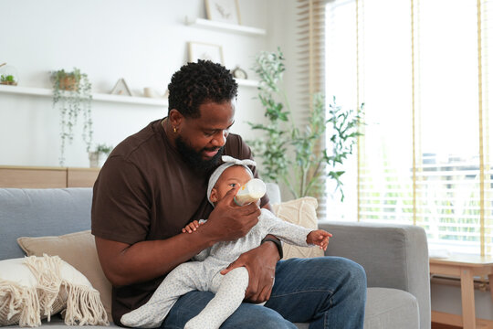 Tender moment as a loving African American father holds and bottle-feeds his infant daughter while sitting on a cozy sofa in a bright living room.