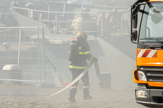 Firefighter in action during an emergency rescue at a port. A brave professional in uniform holding a fire hose with heavy smoke, a ship, and a fire truck in the background.