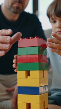 Vertical video: Grabbing red block, dad navy tee, son blue polo placing it building tower at home