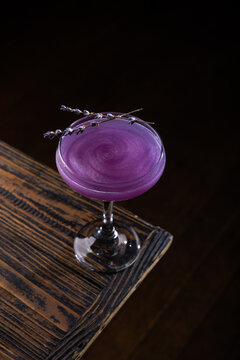 Purple cocktail served in a glass on a wooden table in a dimly lit bar setting at night