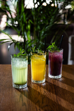 Colorful drinks sit on a wooden table in a lively bar setting with green plants in the background during evening hours