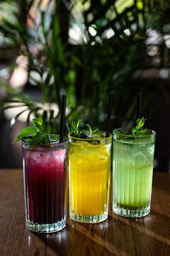 Colorful drinks sit on a table with green plants in the background during a bright afternoon at a cafe