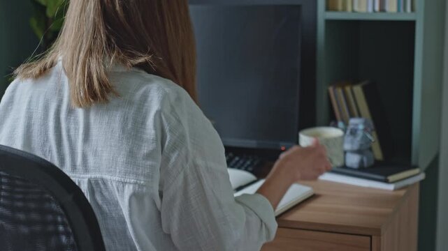 Woman writing in notebook and drinking coffee at desk in home office. Concept of remote work, planning and note taking