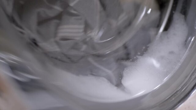 Wet laundry spins inside the drum of a front-loading washing machine. White soapy foam and bubbles swirl against the glass door during a cleaning cycle. The fabric moves in a rhythmic motion