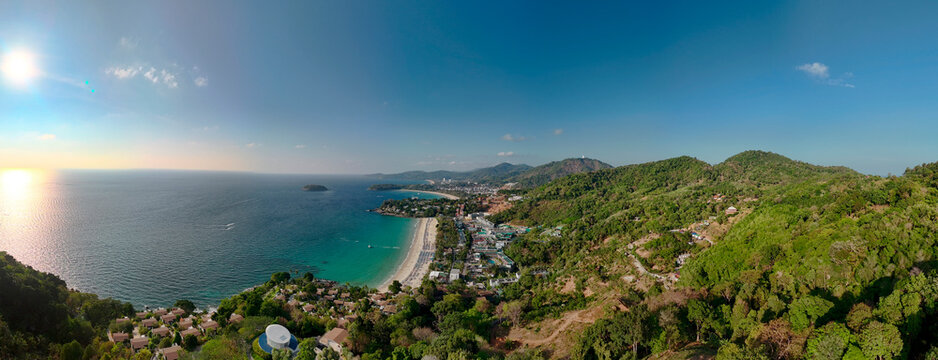Aerial view shows a long Kata beach in Phuket with clear blue water and white sand. Green hills rise behind the beach and many hotels are visible near the shoreline.