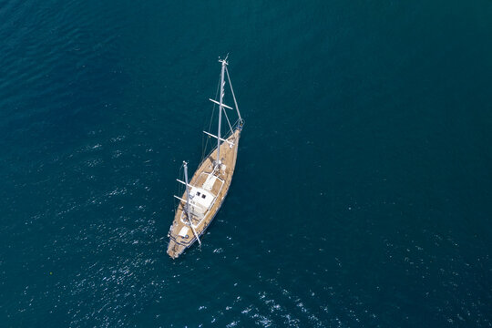 From an aerial perspective, a classic wooden ketch gracefully sails across the shimmering, tranquil sea on a bright, peaceful day